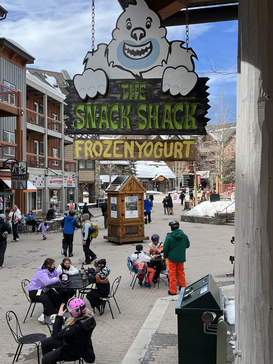 The Snack Shack sign with the yeti over River Run Village in winter, skiers at the tables below